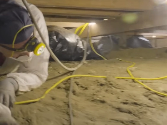 Technician in protective gear lying in a crawl space with lights and yellow cords over bare soil