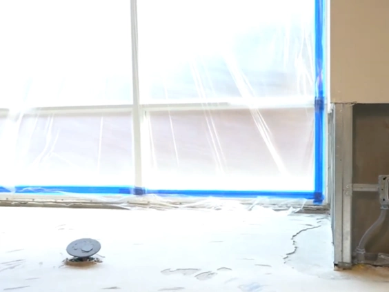 Large window covered in plastic sheeting taped with blue tape above a floor with dust residue