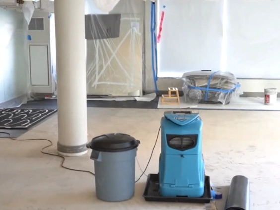 Large room with furniture wrapped in plastic and taped, a blue dehumidifier on a tray and a trash can on the floor