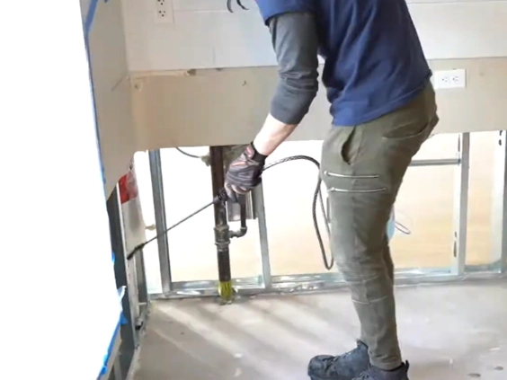 Worker using a hand sprayer on an open wall cavity with metal studs and a pipe below a counter