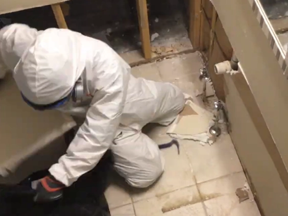 Technician in a white suit and respirator kneeling in a bathroom with the wall opened up beside the tub and floor tile