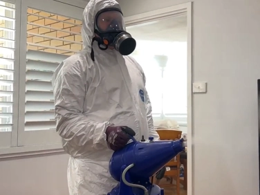 Technician in a protective suit and respirator holding a blue sprayer in a room with window shutters