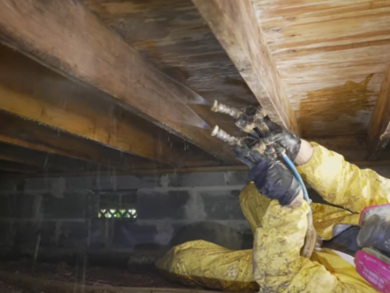 Technician in a yellow protective suit working on floor joists above a block foundation wall in a crawl space