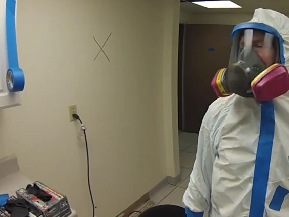 Technician in a white suit with blue tape and a full face respirator standing in a room